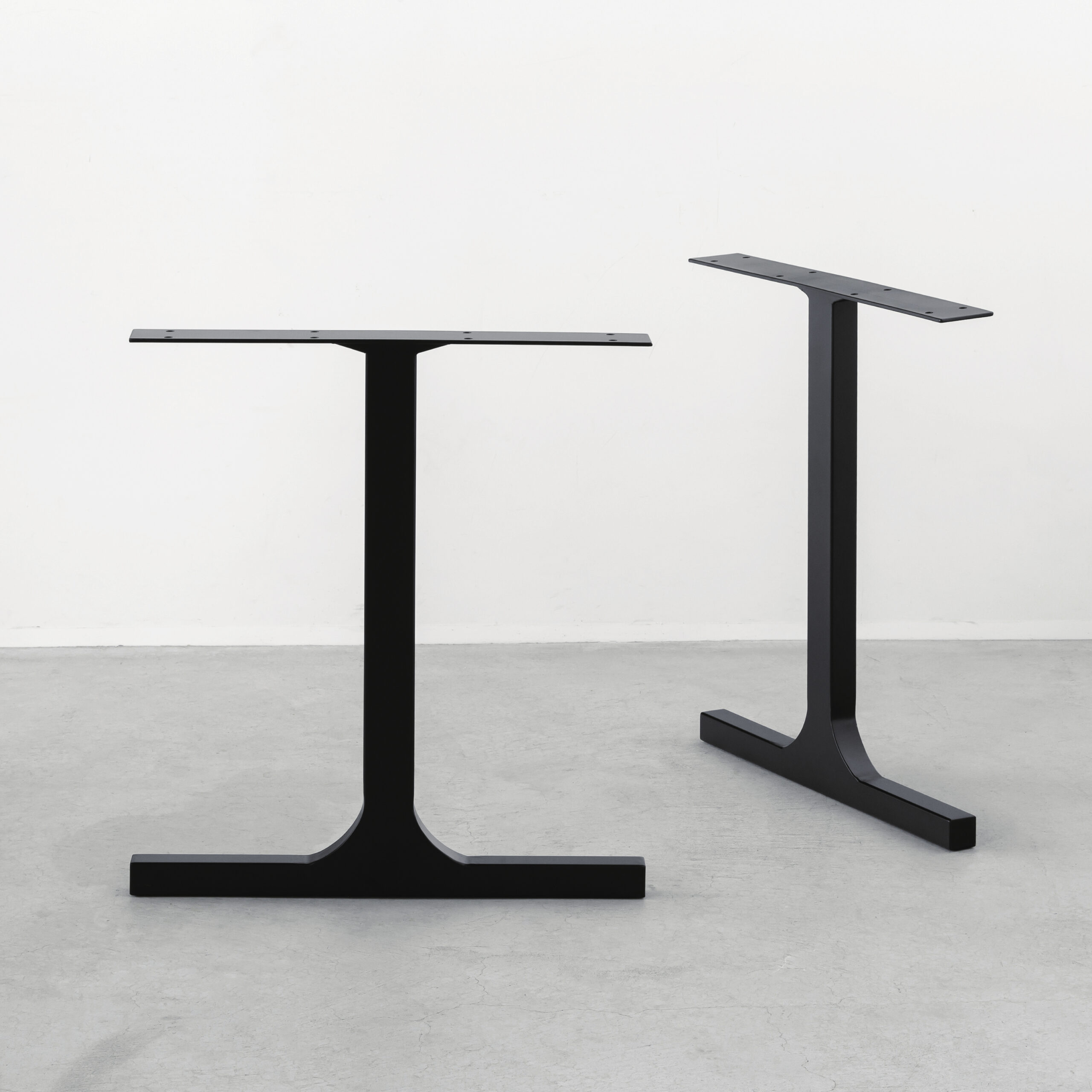 Two black metal table legs with horizontal tops against a pale wall and concrete floor.