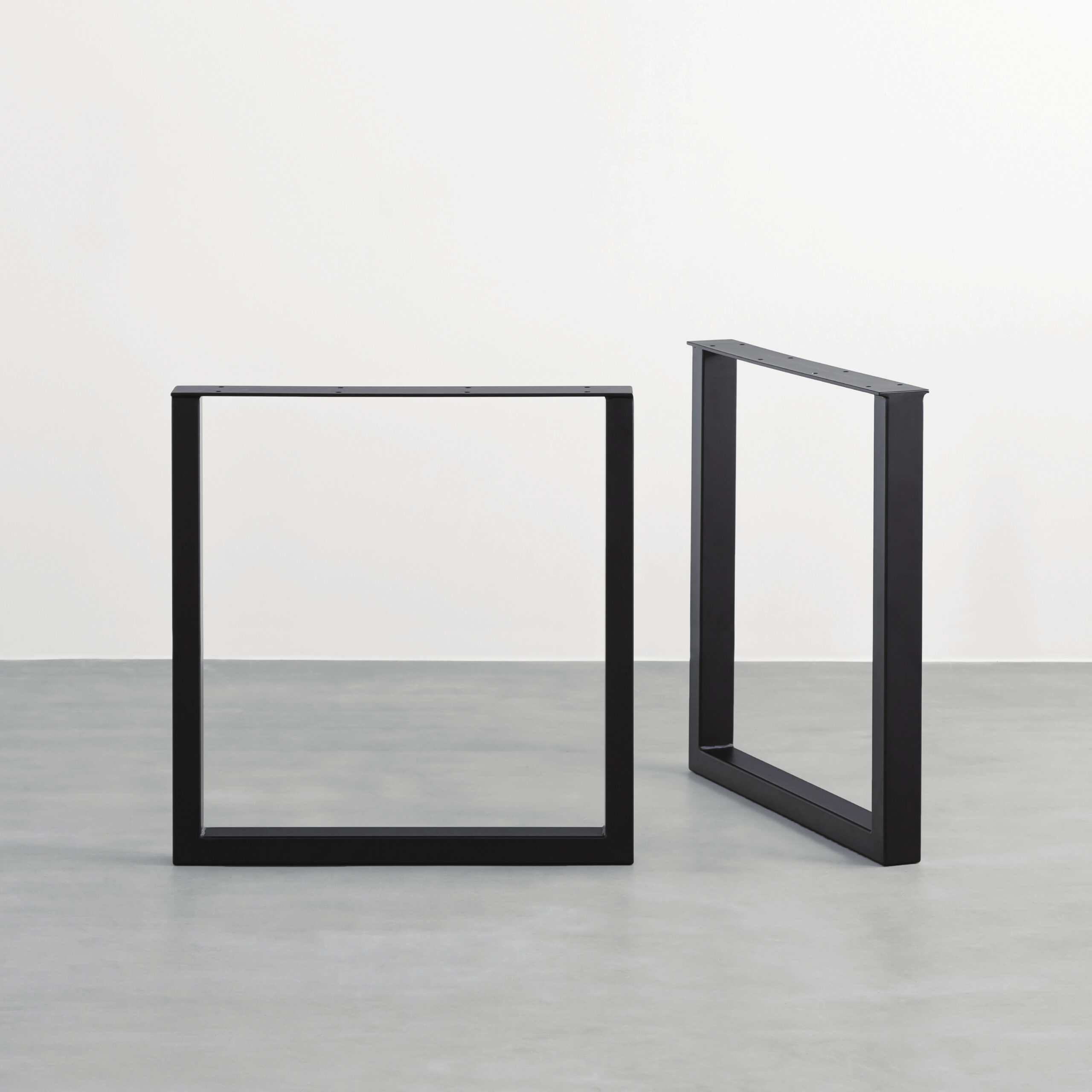 Two black metal rectangular frames standing on a pale floor near a white wall, resembling minimalist sculpture elements.
